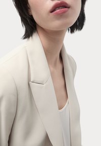 Blazer su misura beige chiaro con spalline e rever a punta, indossato sopra un top bianco. Tessuto liscio, linee pulite e vestibilità moderna.