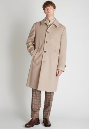 Homme debout avec une main dans la poche, portant un manteau beige, un pull zippé, un pantalon à carreaux et des bottes en cuir marron sur un fond uni.