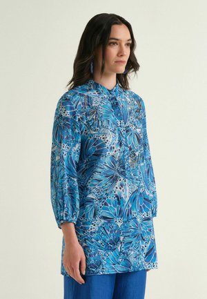 Woman wearing blue butterfly-print long-sleeve blouse with high collar and matching blue earrings, standing against plain light background.