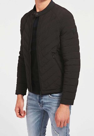 Mann trägt schwarze gesteppte Jacke mit Reißverschluss und Druckknopfkragen sowie hellblaue, abgenutzte Jeans und steht vor einem weißen Hintergrund.