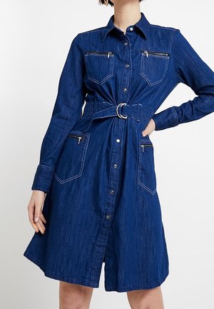 Robe chemise en denim bleu foncé avec un col, des manches longues, deux poches poitrine avec zippers et une taille cintrée mise en valeur par une ceinture.