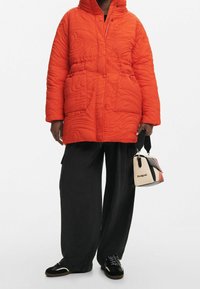 Chaqueta acolchada naranja con capucha y botones a presión, combinada con pantalones negros holgados y un pequeño bolso multicolor. Incluye zapatos negros.