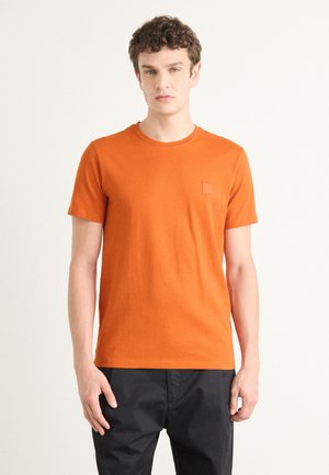 Oransje bomulls t-skjorte med rund hals og korte ermer. Har et lite teksturert logo-patch på venstre bryst. Enkel design.