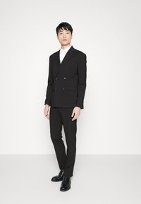 Chaqueta de traje negra de corte cruzado con botones en contraste y pantalones ajustados, combinada con una camisa blanca de cuello y zapatos negros.