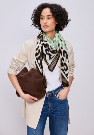 Femme aux cheveux bouclés portant une écharpe imprimé léopard, une veste beige, une chemise blanche, un jean bleu, tenant une grande pochette en suède marron.