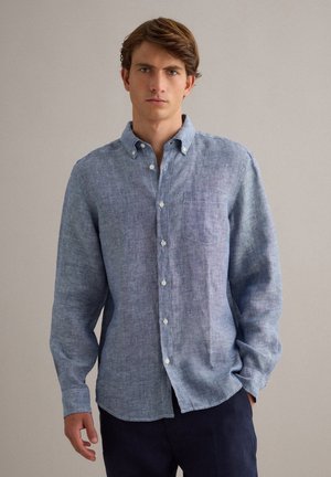 Jonge man draagt een lichtblauw linnen overhemd met knopen en een borstzak en donkere broek, staand tegen een effen achtergrond.