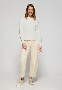 Cremefarbener Pullover mit geripptem Saum, kombiniert mit hellbeigen, weiten Hosen. Das Modell trägt beiger Sneakers und hat lange Haare. Einfache, lockere Passform.