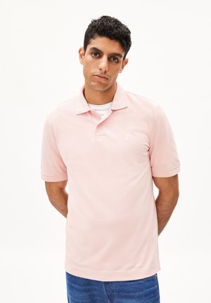 Polohemd aus weichem pinken Stoff mit kurzen Ärmeln und Kragen, versehen mit einem dezenten Logo auf der linken Brust, kombiniert mit blauen Jeans.