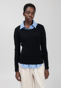 Zwarte trui met lange mouwen en een geribbelde zoom, gelaagd over een lichtblauw gestreept shirt. Het materiaal lijkt glad, met een aansluitend ontwerp.