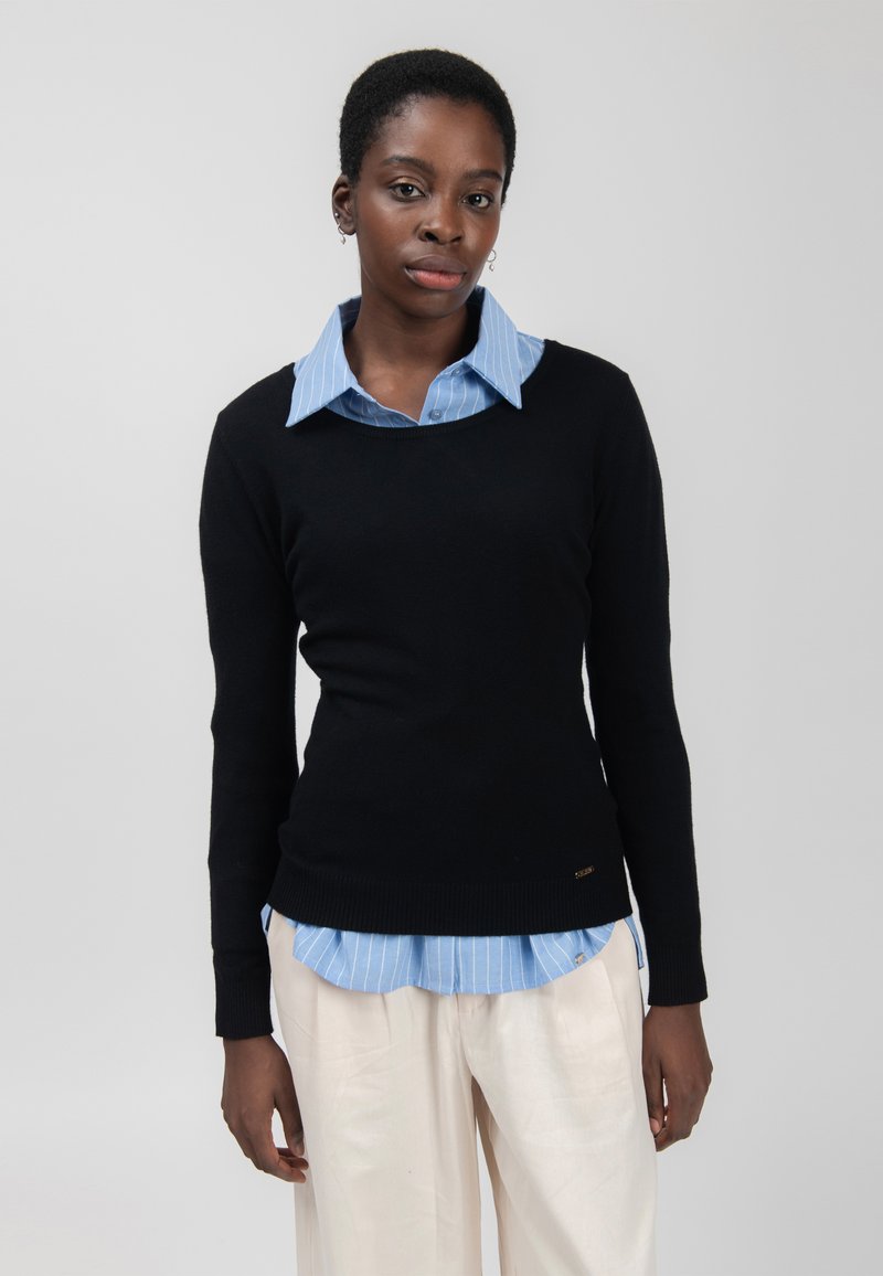 Zwarte trui met lange mouwen en een geribbelde zoom, gelaagd over een lichtblauw gestreept shirt. Het materiaal lijkt glad, met een aansluitend ontwerp.