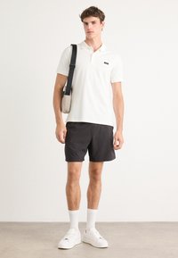 Balti polo marškiniai su trumpomis rankovėmis, juodas logotipas, derinami su juodais šortais. Aksesuarai apima smėlio spalvos kryžminį maišelį ir baltus sportbačius.