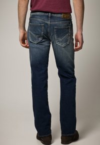 Jeans de mezclilla azul oscuro con un corte recto, que presentan bolsillos traseros con detalles de costura y una etiqueta de cuero marrón en la cintura.