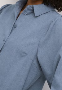 Chemise boutonnée bleu clair texturée avec un col classique et des manches légèrement bouffantes, dotée de deux poches avant et d'un tissu lisse.