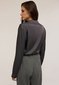 Donna con lunghi capelli castani indossa una blusa di raso grigio scuro e pantaloni grigi a righe, volta verso sinistra su uno sfondo uniforme.