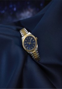 Ceas de mână Timex argintiu și auriu cu un cadran albastru, marcaje aurii pentru ore și o brățară metalică bicoloră pe un fundal de țesătură albastră închisă.