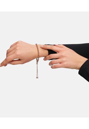 Bracelet en chaîne en bronze avec un détail de pampille, comportant des maillons ronds et un fermoir. Porté sur un poignet contre une manche noire.