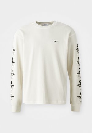 Dolgi rokavi, off-white majica iz vaflja z ribanim ovratnikom in črnimi grafičnimi vzorci na rokavih. Ima majhen logo na prsih.