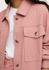 Jasnoróżowa kurtka wykonana z teksturowanej tkaniny, z kołnierzem, dwoma kieszeniami na piersi i okrągłymi guzikami. Krój charakteryzuje się luźnym fasonem i mankietami na rękawach.