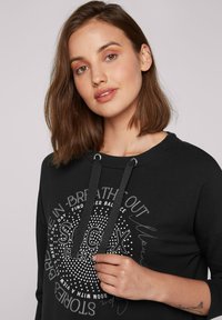 Sudadera negra con un gráfico redondo en texto blanco y puntos. Cuenta con capucha ajustable y una textura suave. Los detalles incluyen frases motivacionales impresas.