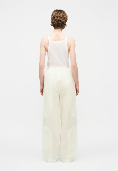 Personne aux cheveux courts et bouclés, portant un haut blanc sans manches et un pantalon ample à jambes larges de couleur blanc cassé, debout de dos sur un fond blanc.