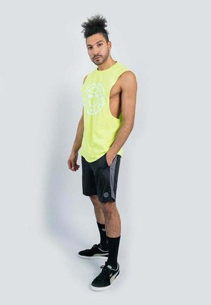 Mann mit lockigem Haar, der ein neongelbes ärmelloses Shirt, schwarze Sportshorts, schwarze mittellange Socken und schwarze Turnschuhe trägt, steht vor hellem Hintergrund.