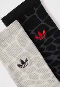 Δύο ζευγάρια κάλτσες: το ένα μαύρο με υφασμένο μοτίβο και κόκκινο λογότυπο, το άλλο ανοιχτό γκρι με παρόμοιο μοτίβο και μαύρο λογότυπο.