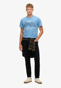 Lys blå meleret t-shirt med sort vintage tryk, sorte jeans og en ternet skjorte knyttet om taljen; cremefarvede sneakers med brune detaljer.