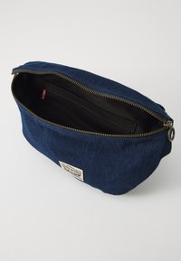 Denim torbica z temno modrim zunanjim delom, zlatim zadrgo in bež oznako. Notranjost je obložena s črnim materialom in ima majhen žep za organizacijo.