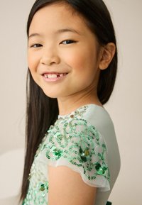 Šviesiai turkio spalvos suknelė, kurios trumpi, plazdantys rankovės yra papuoštos žaliomis blizgučiais ir perlais. Minkštas audinys šviesiame fone.