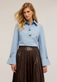 Camicia azzurro chiaro a maniche lunghe con colletto e grandi bottoni, abbinata a una gonna in pelle marrone plissettata con dettagli in bottoni argentati.
