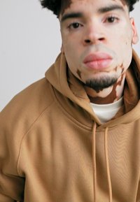 Brown hoodie with a textured finish, featuring a hood and drawstrings, worn above a light-colored t-shirt. Close-up of a person’s face.