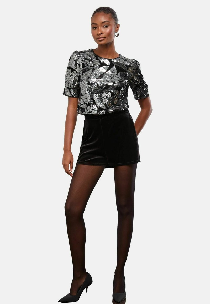 Čierne zamatové šortky v kombinácii s lesklým, flitrovým topom s kvetinovým vzorom. Modelka nosí priesvitné čierne pančuchy a špicaté čierne podpätky.