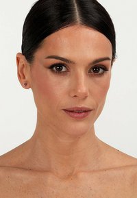 Model met een gemiddelde huidtoon, donker haar dat naar achteren is gestyled, draagt kleine stud oorbellen. Netjes opgemaakt met een focus op de ogen en lippen. Gladde huidtextuur.