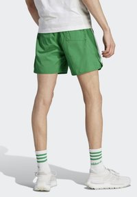 Green swim shorts with a loose fit, side pockets, and a back pocket, made from lightweight material. Paired with white shoes and striped socks.