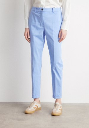 Personne portant un pantalon bleu clair sur mesure coupé à la cheville, une chemise blanche à manches longues et des baskets beiges avec semelles en gomme, debout sur un sol gris.
