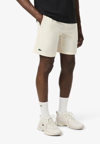 Pantalones cortos deportivos color crema con bolsillos y logo de Lacoste en verde. Combinados con calcetines blancos y calzado ligero. Material suave y liviano.