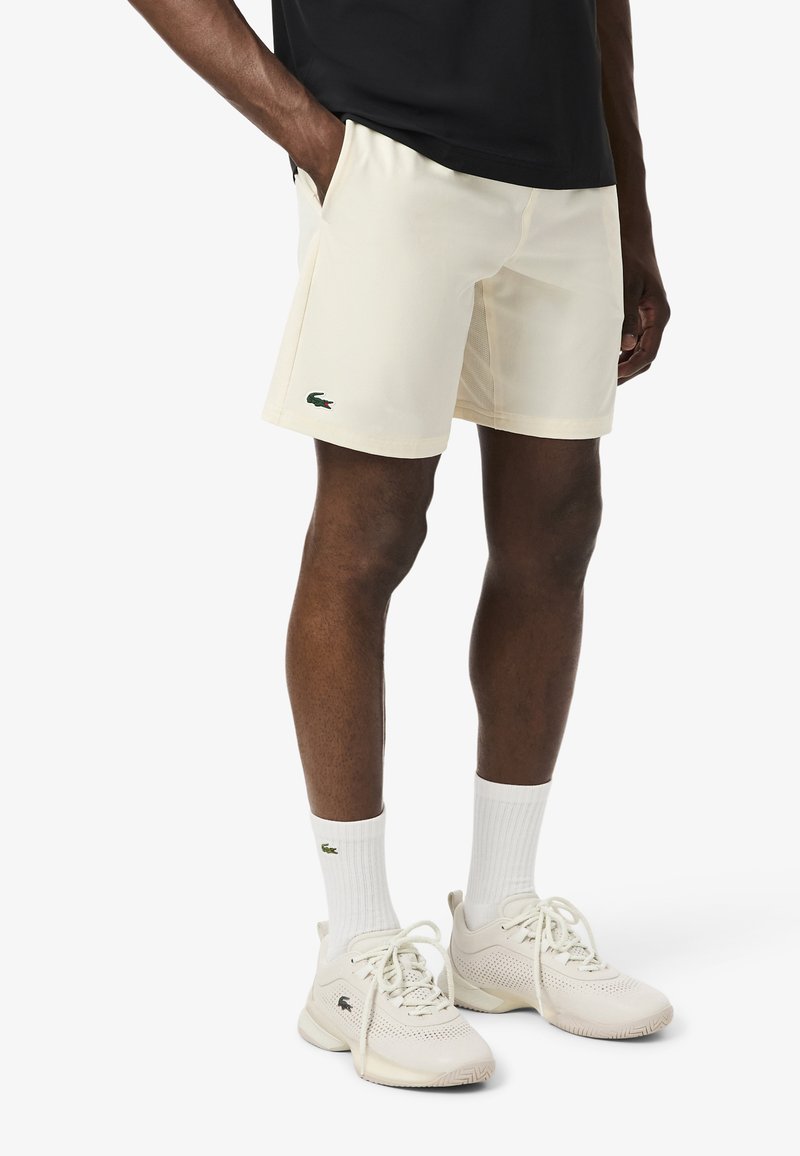 Pantalones cortos deportivos color crema con bolsillos y logo de Lacoste en verde. Combinados con calcetines blancos y calzado ligero. Material suave y liviano.
