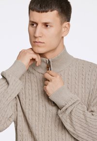 Beżowy sweter dziany z wysokim kołnierzem i zapięciem na zamek, z teksturą w warkocz i ściągaczami przy rękawach.