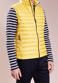Gilet matelassé jaune avec col montant, quilting vertical et poches zippées. Porté par-dessus une chemise à rayures navy et blanches à manches longues.
