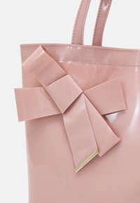 Borsa tote rosa lucido con un grande fiocco realizzato nello stesso materiale. Presenta bordi cuciti e un'etichetta accentuata in oro sulla parte inferiore.