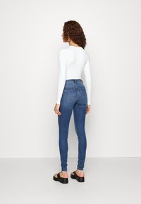 Jeans de ganga azuis de cintura alta, ajustados ao corpo, com desbotamento mínimo e dois bolsos traseiros, combinados com uma blusa de mangas compridas branca e sandálias pretas.