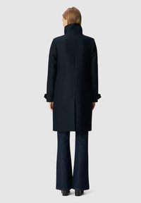 Abrigo de lana negro con cuello alto, corte recto y un sutil detalle en los hombros. Llevado con jeans acampanados oscuros y tacones gruesos negros.