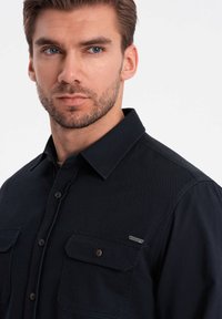 Camisa azul oscura de algodón, con cuello abotonado, dos bolsillos en el pecho y un pequeño logotipo metálico en el bolsillo izquierdo.