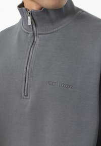 Grå kvart-zip hettegenser med ribbestrikket krage og preget logo. Glatt tekstur og uformell design, med sølvfarget glidelåsdetalj.