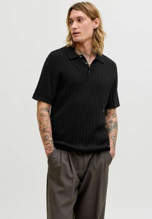 Mann mit schulterlangem Haar und Tätowierungen trägt ein schwarzes, kurzärmeliges Strick-Poloshirt und lockere braune Hosen, steht mit den Händen in den Taschen.