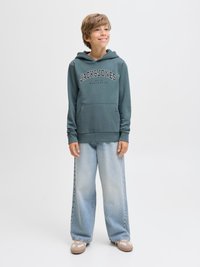 Hanorac cu glugă verde-albăstru închis, cu buzunar frontal și logo-ul „JACK&JONES”, asortat cu blugi largi albastru deschis și adidași maro.