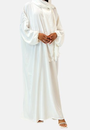 Kobieta ubrana w lejącą się białą abaję z długimi bufiastymi rękawami i pasującymi białymi sandałami, stojąca z delikatnie splecionymi rękami.