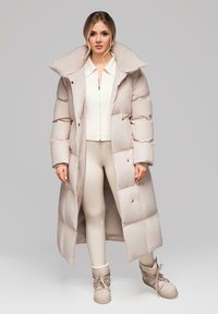 Žena v dlouhém béžovém prošívaném kabátu, krémové zipové mikině, odpovídajících legínách a béžových zimních botách s tkaničkami stojí proti šedému pozadí.