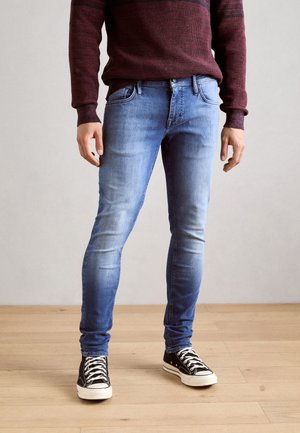 Jeans ajustados de mezclilla azul con desvanecimientos, diseño de cinco bolsillos, presillas para cinturón, y combinados con zapatillas de lona negras con cordones blancos.