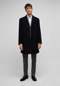 Marineblauer Wollmischungs-Mantel mit drei großen Knöpfen, seitlichen Taschen und Revers, kombiniert mit grauen Karohosen und schwarzen Lederstiefeln.